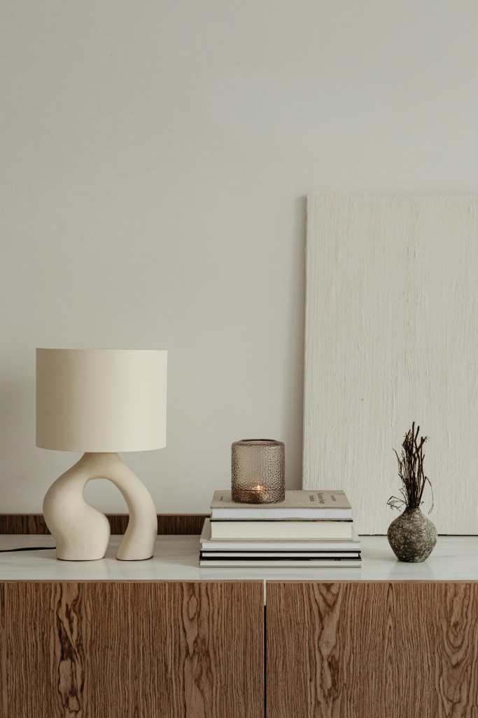 Neutral minimalist home decor featuring a beige table lamp, stacked books, and simple ceramic accessories on a wooden cabinet.
