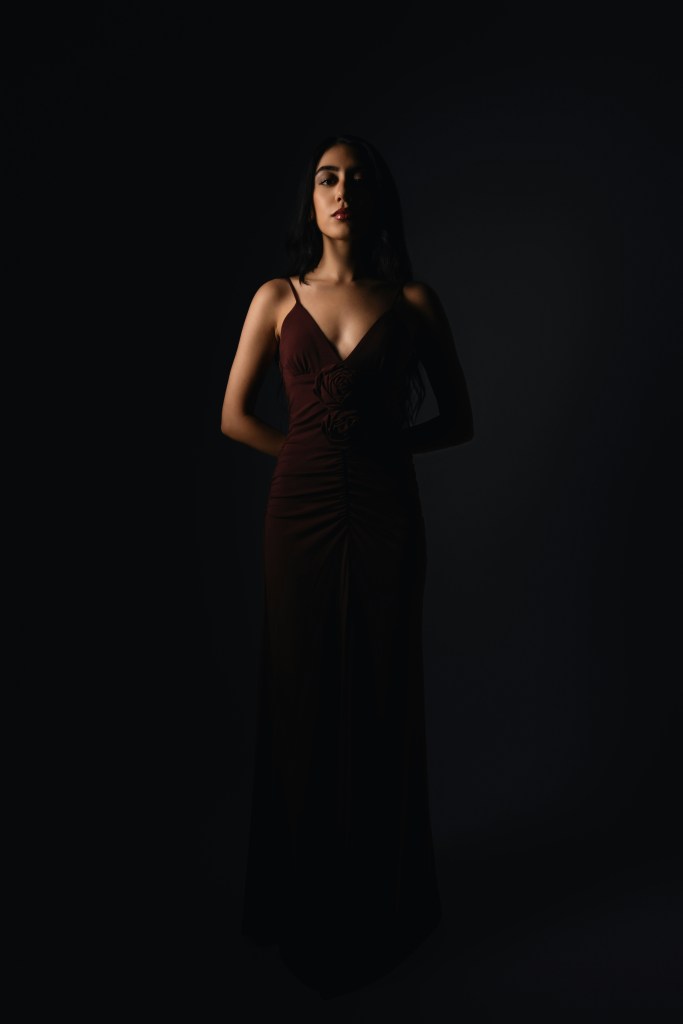 Woman in a deep-toned floor-length gown standing in dramatic lighting, representing minimalist winter eveningwear.