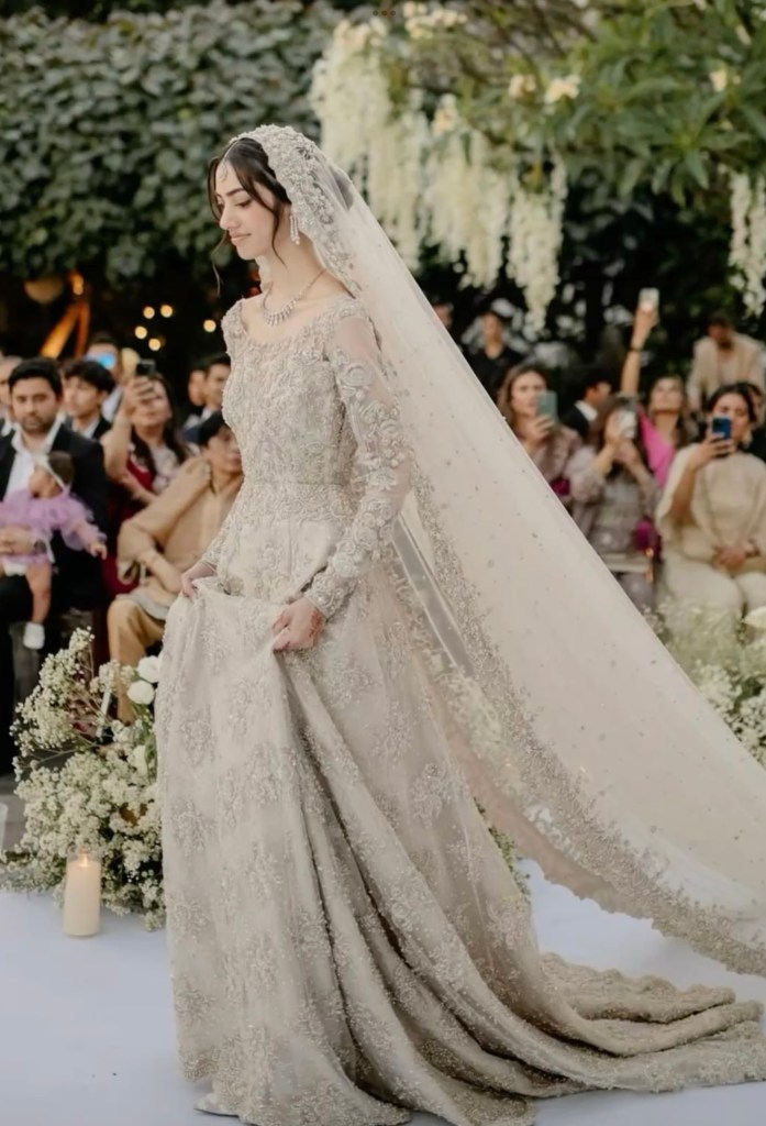 Bride wearing a long ivory embroidered gown with full sleeves and a flowing veil, walking outdoors during a wedding ceremony.
