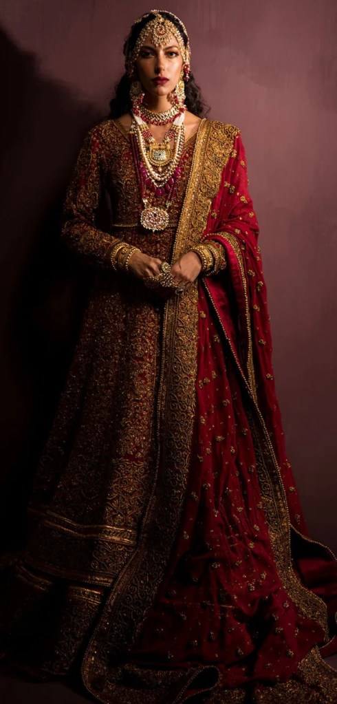 Red Mughal-inspired bridal outfit with heavy zardozi embroidery and layered gold jewelry.