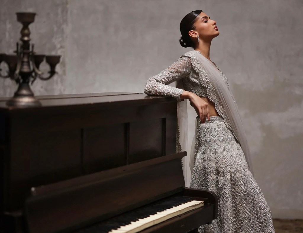 Model wearing a silver beadwork lehenga with sheer dupatta, leaning against a piano in a modern editorial bridal setting.