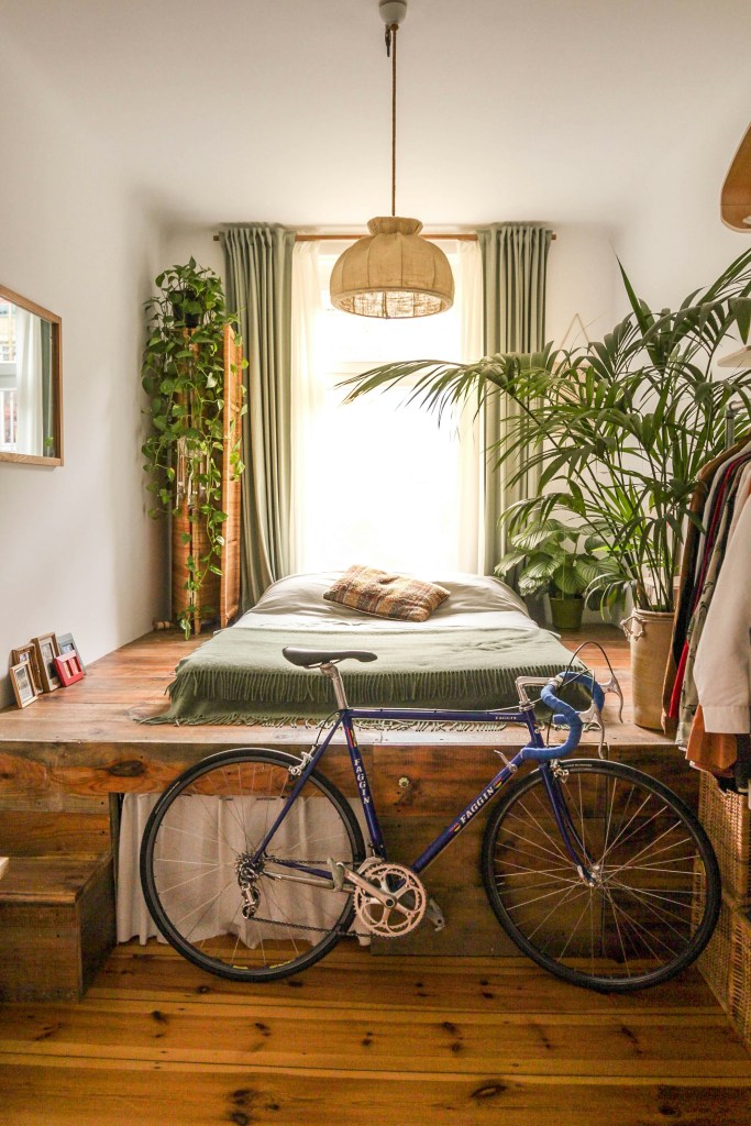 A cozy bedroom featuring a bike beside a raised wooden bed, with green curtains and plants near the window, creating a warm and inviting atmosphere.