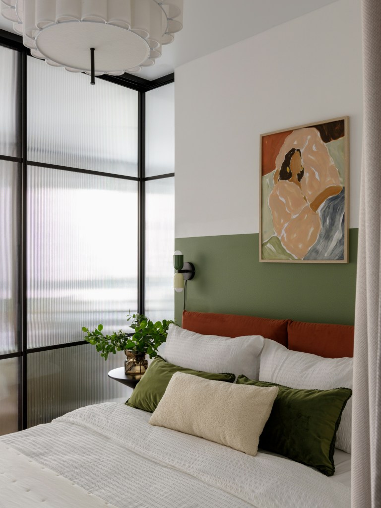 A cozy bedroom featuring a bed with white and green pillows, a green accent wall, and a piece of abstract art hanging above. There is a stylish ceiling fixture and large windows providing natural light, along with a green plant on a small side table.
