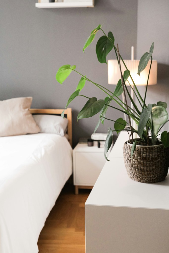 A cozy bedroom scene featuring a white bed and a bedside table with a woven plant pot and decorative items. A lamp emits warm light against a grey wall.