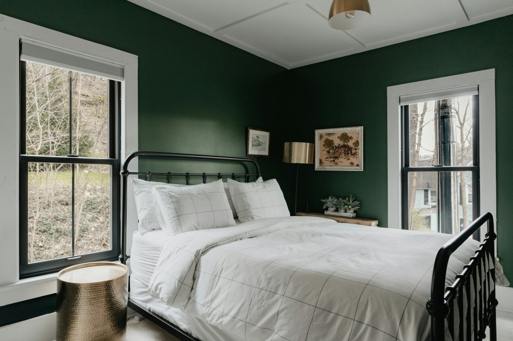 A cozy bedroom featuring a black metal bed frame with white linens, dark green walls, large windows with black frames, and a bedside table with a decorative plant.