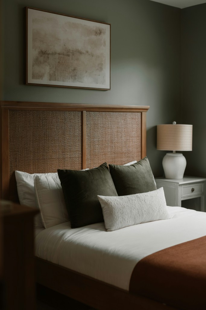 A beautifully designed bedroom featuring a wooden headboard with a woven texture, a neatly made bed with white linens, dark green accent pillows, and a decorative beige pillow. The room has a soft green wall and a piece of abstract wall art above the bed. There is a bedside table with a lamp on the side.