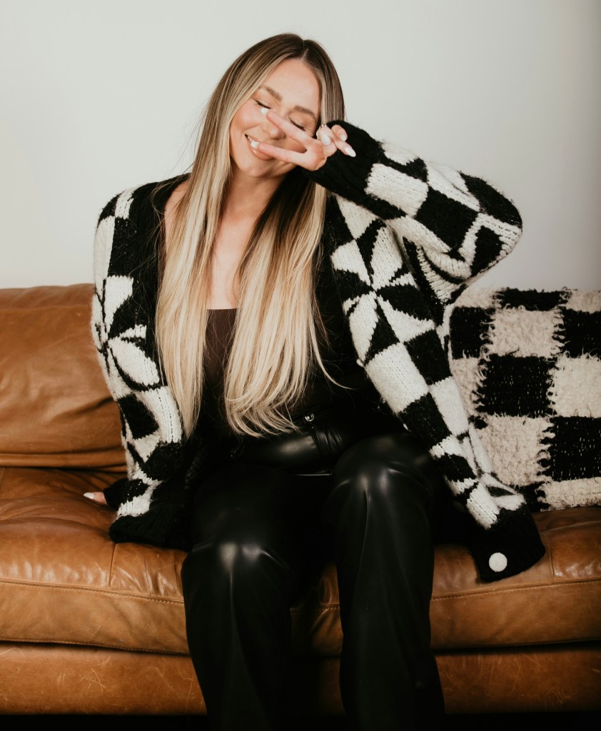 Woman in a chunky black and white check cardigan and black leather trousers, seated on a tan leather sofa