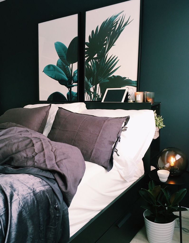 A cozy bedroom featuring a neatly made bed with dark gray bedding and pillows, decorative plant-themed artwork on the wall, and a stylish lamp on a bedside table.